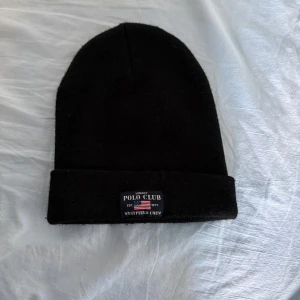 Svart mössa Polo Club - Svart, klassisk mössa från Polo Club med uppvikt kant och en liten etikett framtill med logga och amerikansk flagga. Perfekt accessoar för kyliga dagar och enkel att matcha med olika outfits.