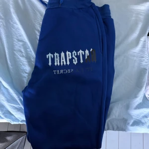 Blå mjukisbyxor Trapstar M - Snygga blå mjukisbyxor från Trapstar med tryck på benet i vitt och guld. Byxorna har ribbade muddar vid benslut och midja samt mjukt fleecefoder på insidan. Perfekt för chill eller streetwear-look.