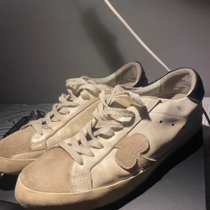 The Gallerist vita sneakers med klubbmärke - Unika sneakers från The Gallerist i vit skinn med beige mockadetaljer och svart häl. Klubbmärke i mocka på sidan och leopardmönstrad rem bak. Platt sula och snörning framtill. Perfekt för dig som gillar detaljer och streetstyle.