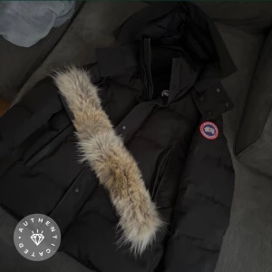 Svart dunjacka från Canada Goose - Säljer en svart dunjacka från Canada Goose med avtagbar huva och pälsdetalj. Jackan har klassisk patch på ärmen, dragkedja och knappar framtill samt två stora fickor. Perfekt för kalla vinterdagar och riktigt snygg streetstil. Kostar för tillfälligt 16,800 kr