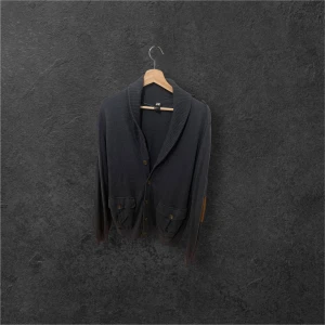 Mörkgrå cardigan med bruna detaljer H&M - Snygg mörkgrå kofta från H&M med ribbad krage och bruna mockadetaljer på armbågarna. Framtill finns två fickor med knappar och koftan stängs med knappar. Det är storlek M 