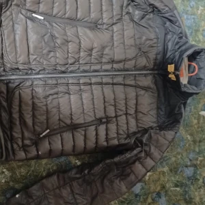 Grå Parajumpers pufferjacka herr - Snygg grå pufferjacka från Parajumpers med quiltad design och hög krage. Jackan har dragkedja framtill, två snedställda fickor med dragkedja och en patch på ärmen. Insidan är ljus och jackan har coola detaljer som orange hängare och Parajumpers-logga.