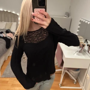 Svart blus med spets och volanger - Supersnygg svart blus med transparent spets upptill och blommigt mönster. Långa ärmar och volangdetaljer längs axlar och sidor ger en extra cool vibe. Perfekt att styla med jeans eller kjol för en trendig look.