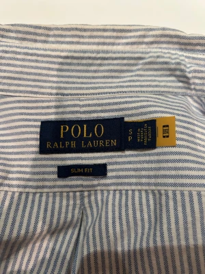 Randig skjorta från Polo Ralph Lauren - Slim fit skjorta från Polo Ralph Lauren i blått och vitt med klassiska vertikala ränder. Skjortan har lång ärm, knappar framtill och broderad logga på bröstet. Tillverkad i mjuk bomull, perfekt för en clean och fräsch look.