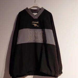 Svart Arsenal F.C. tröja med snörning - Cool svart tröja från Arsenal F.C. med grå detaljer och broderad logga samt årtalet 1886 på bröstet. V-ringad hals och långärmad modell med snörning i nederkant för extra stilpoäng. Perfekt för dig som älskar fotboll och vill sticka ut. Väldigt unik eftersom den inte heller finns att köpa ny i Sverige.