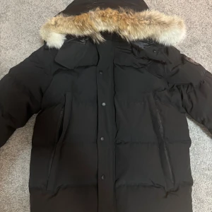 Canada goose - Säljer en svart dunjacka med fluffig pälsdetalj på huvan. Jackan har långa ärmar, stora fickor med dragkedja och knappar framtill. Perfekt för kalla dagar och har en riktigt schysst oversized look.