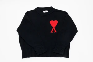 Svart stickad tröja från Ami Paris  - Svart stickad tröja från Ami med rund hals och ribbade muddar. På bröstet finns ett stort rött hjärta med bokstaven A. Tröjan har lång ärm och är tillverkad i mjuk ull, perfekt för kyliga dagar. Snygg och stilren design som sticker ut. Ett litet streck precis ovanför loggan syns vid noga inspektion.