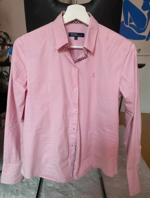 Rosa randig skjorta från Polo Ralph Lauren - Snygg rosa skjorta från Polo Ralph Lauren med tunna vita ränder och broderad logga på bröstet. Insidan av kragen har ett rutigt mönster i rött, vitt och blått. Skjortan har klassisk krage, knappar framtill och långa ärmar. Tillverkad i mjuk bomull.