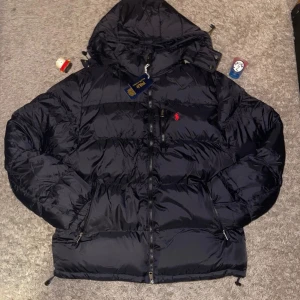 Svart pufferjacka från Polo Ralph Lauren - Svart pufferjacka från Polo Ralph Lauren med huva och flera dragkedjor. Jackan har det klassiska röda Polo-logot broderat på bröstet och är tillverkad i glansig polyester. Perfekt för kalla dagar med sin tjocka och vadderade design. Vi kan prata om priset!