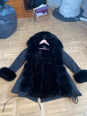 Svart parkajacka med pälsdetaljer - Säljer en svart parkajacka med fluffiga pälsdetaljer på huva och ärmslut. Jackan har snörning i midjan och nederkant, samt långärmad design. Insidan är fodrad med mjuk päls för extra värme och stil. Storlek XS/S men passar även någon som har M (små i storleken) Perfekt för dig som vill ha en cool och lyxig vinterlook.