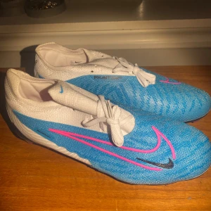Nike Phantom GX blå fotbollsskor - Nike Phantom GX fotbollsskor i blått och vitt med rosa och svart swoosh-detaljer. Ovandel i syntetmaterial med snörning och rund tå. Sulan har rosa dobbar och coola blå detaljer för extra grepp på planen. Perfekta för dig som vill sticka ut på matchen.