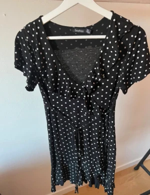 Prickig kortklänning från Boohoo - Supersöt kortklänning från Boohoo med vita prickar. Klänningen har volangdetaljer vid halsringningen, v-ringad front och knytband i midjan. Materialet är mjukt och lätt, perfekt för varma dagar. Kort arm, figurnära passform och stretch. Gula klänningen visar passformen.