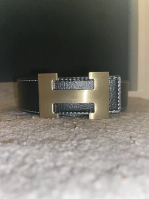 Svart skärp med H-spänne - Säljer ett svart med ett ikoniskt H-format spänne i guldtonad metal med vit kontrastsöm längs kanterna. Perfekt accessoar för att lyfta din outfit med en lyxig touch.