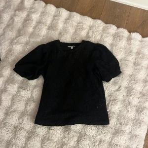 Svart blus med puffärm från Object - Svart blus från Object i storlek XS med puffiga korta ärmar och broderade detaljer över hela plagget. Blusen har ett snyggt hålmönster och rund halsringning. Perfekt för dig som gillar coola och kreativa detaljer i din stil.