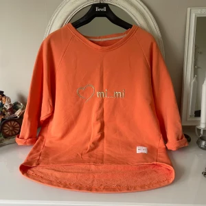  mi...mi sweatshirt  - Cool orange sweatshirt från mi...mi med broderats hjärta och text på bröstet. Tröjan har rund hals, trekvartsärmar och är lite längre baktill. Tillverkad i mjuk bomull med lite stretch för extra komfort. Perfekt för dig som gillar streetstyle och färg.