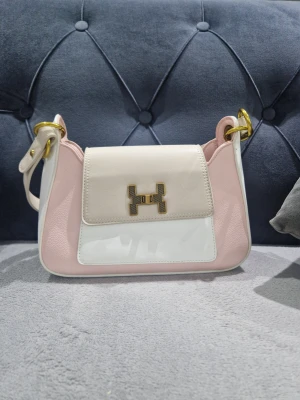 Vit och rosa axelväska med H-spänne - Snygg axelväska i vitt och ljusrosa med guldfärgat H-spänne framtill. Väskan har en mjuk, rundad form och detaljer i guld på remmarna. Materialet är skinnimitation och väskan har en stilren look som sticker ut med sin färgkombination.