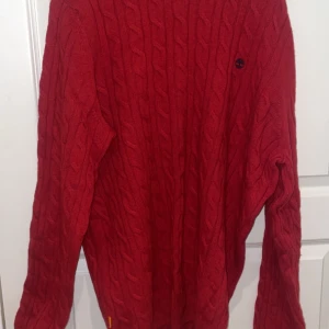 Röd kabelstickad Timberland tröja XL - Snygg röd kabelstickad tröja från Timberland i lamullsmix. Tröjan har rund hals, lång ärm och ett broderat Timberland-logo på bröstet. Perfekt för dig som gillar klassisk stil med en modern twist.