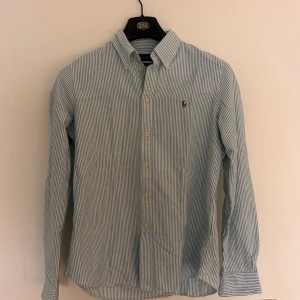 Blårandig skjorta från Ralph Lauren - Snygg blårandig skjorta från Ralph Lauren med klassisk button-down krage och broderad logga på bröstet. Custom fit-modell i mjuk bomull, perfekt för dig som gillar stilrena och tidlösa plagg. Långa ärmar och knappar framtill.
