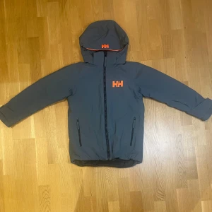 Skidjacka från Helly Hansen i storlek 140 - Snygg mörkgrå skidjacka från Helly Hansen, perfekt för skidsäsongen. Jackan har en orange HH-logga på bröstet och huvan. Jackan har avtagbar huva, två dragkedjeförsedda fickor och justerbara ärmslut med kardborre. Insidan är svart och materialet är slitstark polyester. Jackan har även en liftkorthållare på vänster arm. Pris kan diskuteras!
