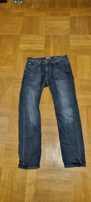 Blå jeans från Dressmann Regular Fit - Snygga blå jeans från Dressmann i modellen Regular Fit. Klassisk femficksdesign med raka ben och normal passform. Jeansen har en mörk tvätt med lätt slitning och är tillverkade i slitstarkt denimtyg. Perfekt för dig som gillar enkel och stilren look. Originalpris: 800