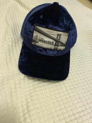 Blå sammetstruckerkeps Hatstore - Snygg blå truckerkeps från Hatstore med framsida i glansig sammet och mesh baktill. Kepsen har ett svartvitt tryck med New Yorks skyline och bro på fronten. Justerbar passform och unik look som sticker ut.