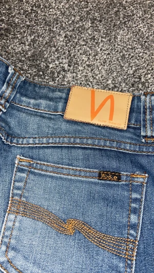 Blå jeans från nudie  - Säljer ett par blå  jeans från Nudie med klassisk orange söm och Nudie-logga på bakfickan. Jeansen har en snygg tvätt med ljusare partier och fem fickor. Perfekt passform för dig som gillar smalare ben.