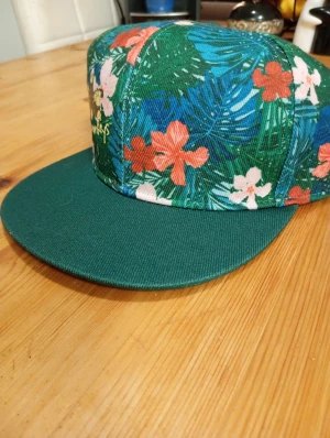 Grön blommig keps med platt skärm - Snygg keps med platt grön skärm och färgglatt blommigt mönster i grönt, blått, rött och vitt. Perfekt för dig som vill sticka ut med tropisk vibe. Kepsen har klassisk form och är tillverkad i bomullstyg.