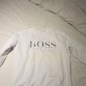 Vit långärmad tröja Hugo Boss - Snygg vit långärmad tröja från Hugo Boss med silverfärgat BOSS-tryck på bröstet. Tröjan har en klassisk rund hals och är gjord i mjuk bomull, perfekt för en clean och stilren look. Passar dig som gillar enkla men lyxiga plagg.