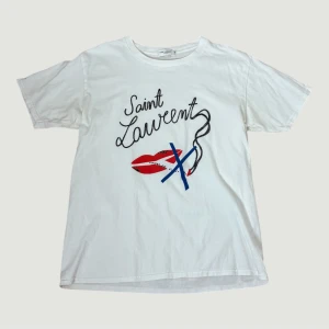 Sjukt snygg Saint Laurent tshirt  - Sjukt cool t-shirt från Saint Laurent med coolt grafiskt tryck framtill, där texten 'Saint Laurent' kombineras med röda läppar och blå detaljer. Klassisk rund hals och korta ärmar. Mjuk bomullskvalitet som känns skön mot huden. Nästan till ny, då jag inte använt den, har dock en liten fläck nere i högra hörnet på baksidan som man ser på bilden, men den går säkert bort i tvätten! Storlek M. Köpt här på plick så kan ej garantera äktighet