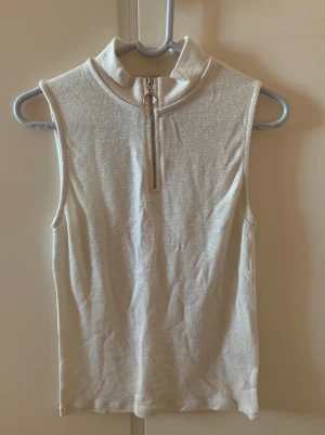 Glittrig polotopp  - Säljer en glittrig beige polotopp från Cubus i storlek M. Toppen är ärmlös och har en hög krage med en snygg silverfärgad dragkedja framtill. Oanvänd med prislapp kvar