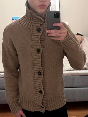 Beige stickad kofta från Zara - Säljer en beige stickad kofta från Zara med hög ribbad krage och stora svarta knappar framtill. Koftan har långärmad passform och breda ribbade muddar vid ärmslut och nederkant. Personligt är perfekt för lager på lager under kyliga dagar. Skick 9/10