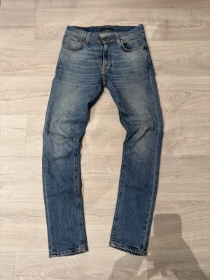 Nudie Jeans - Säljer ett par klassiska blå jeans från Nudie Jeans Co med snygg tvätt och kontrastsömmar. Modellen har fem fickor, normal passform och raka ben. Materialet är slitstarkt jeans i bomull och de har en cool patch med Nudie-loggan bak.