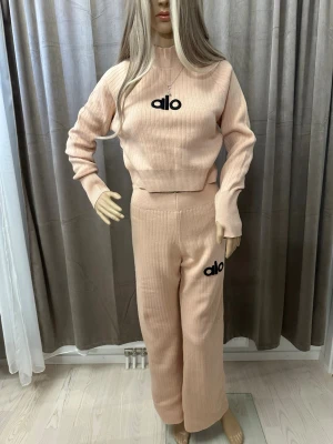 Beige ribbad väst från Alo - Säljer en beige ribbad väst från Alo med svart broderad logga framtill. Västen är stickad i mjukt material och har en hög hals. Perfekt för lager-på-lager och ger en clean look.