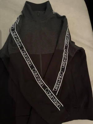 Svart och mörkgrå Calvin Klein ziptröja - Säljer en svart och mörkgrå ziptröja från Calvin Klein med hög krage och hel dragkedja. Tröjan har vita logoband längs ärmarna och ribbade muddar. Perfekt för en chill och sportig look.
