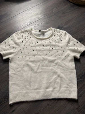 Vit stickad tröja med pärlor - Supersöt vit stickad tröja med korta ärmar och rund hals. Tröjan är dekorerad med pärlor och små metallkulor över axlar och bröst, vilket ger en lyxig och trendig känsla. Perfekt att styla till jeans eller kjol för en chic look.