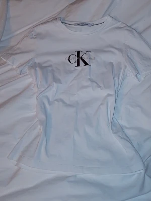 Calvin Klein Jeans t-shirt - Snygg vit t-shirt från Calvin Klein Jeans med stort CK-logotyptryck i vinrött och texten 'Calvin Klein Jeans' över bröstet. Klassisk rund halsringning och korta ärmar. Tillverkad i mjuk bomull för en skön känsla och enkel stil.