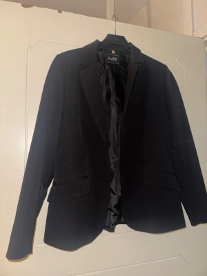 Svart kavaj från Flare Collection - Stilren svart kavaj från Flare Collection i storlek 36. Klassisk modell med krage, två fickor med lock och helsvart foder. Perfekt för dig som vill ha en enkel och snygg kavaj till garderoben.