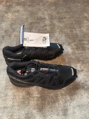 Salomon Speedcross 4 svarta sneakers - Salomon Speedcross 4 sneakers i svart med snörning och robust sula för bra grepp. Tillverkade i slitstark syntet och textil, med logga på sidan och reflekterande detaljer. Perfekta för utomhusaktiviteter och träning.