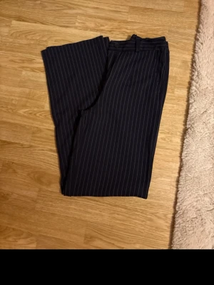 Randiga kostymbyxor från Kappahl - Säljer ett par mörkblå kostymbyxor från Kappahl i storlek 34. Byxorna har smala vita ränder, hög midja och raka, vida ben. Klassisk look med bälteshällor och diskreta fickor. Perfekta för en stilren och trendig outfit.