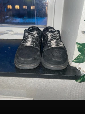 Travis scott Nike air Jordan low 1 - Säljer ett par svarta Nike Air Jordan 1 Lows med vit kontrastsöm och svartvitmönstrade skosnören. Skorna har en klassisk låg siluett, rund tå och är tillverkade i mocka och skinn. Perfekta för dig som gillar streetwear och vill ha en ikonisk sneaker i garderoben.