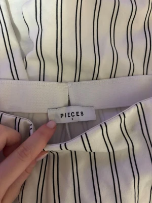 Randiga byxor från Pieces, storlek S - Säljer ett par vita byxor från Pieces i storlek S med tunna svarta vertikala ränder. Byxorna har en rak passform och elastisk midja för extra komfort. Perfekta för dig som gillar en clean och stilren look.