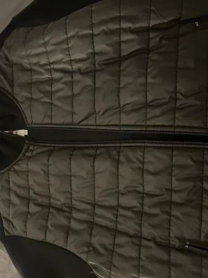 Svart quiltad dunväst från Moncler XL - Säljer en svart quiltad dunväst från Moncler i storlek XL. Västen har en stilren design med vertikala sömmar, dragkedja framtill och diskret Moncler-logga på bröstet. Tillverkad i syntetmaterial och dun, perfekt för lager-på-lager under kyliga dagar.