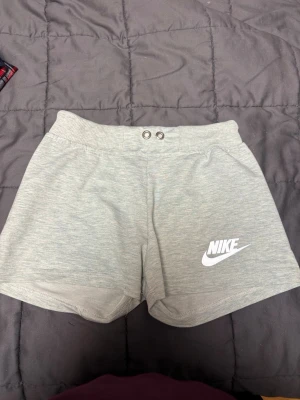 Grå shorts från Nike i bomull - Snygga ljusgrå shorts från Nike med elastisk midja och två metallöljetter framtill. Tillverkade i mjuk bomull och har en vit Nike-logga på benet. Storlek M