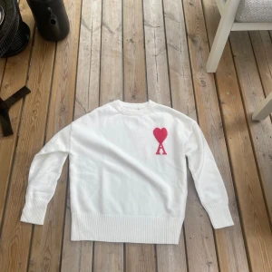 Vit stickad tröja från Ami Paris - Vit stickad tröja från Ami Paris med röd hjärtlogga och bokstaven A på bröstet. Tröjan har rund hals, ribbade muddar och nederkant. Materialet är mjuk bomull. 