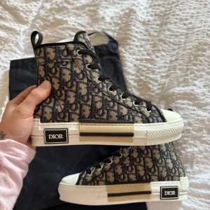 Dior high top sneakers med monogram - Snygga high top sneakers från Dior med ikoniskt monogrammönster i beige och marinblå. Skorna har svart snörning, vit gummisula med Dior-logga och detaljer i svart och beige. Tillverkade i canvas med gummidetaljer och rund tå. Perfekta för dig som vill sticka ut.