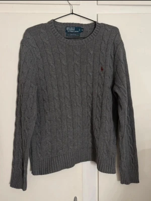Grå kabelstickad tröja Polo Ralph Lauren - Klassisk grå kabelstickad tröja från Polo Ralph Lauren i 100% bomull. Tröjan har rund halsringning, ribbade muddar och den ikoniska röda loggan broderad på bröstet. Perfekt för lager på lager under höst och vinter.