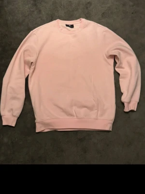 Ljusrosa sweatshirt från H&M - En enkel och stilren ljusrosa sweatshirt från H&M. Tröjan har rund halsringning, långa ärmar och ribbade muddar vid ärmslut och nederkant. Perfekt för en avslappnad look och lätt att matcha med jeans eller mjukisbyxor.