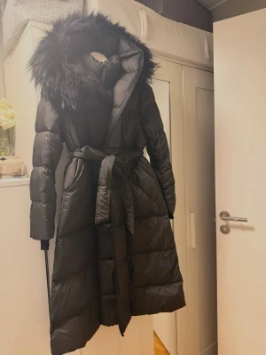 Svart lång pufferjacka med fuskpäls - Säljer en svart lång pufferjacka med stor huva och fuskpälsdetalj. Jackan har bälte i midjan för snygg passform och dragkedja framtill. Perfekt för kalla vinterdagar och riktigt mysig att bära. Materialet är syntet och dunliknande stoppning. Super vaaaarm då min väninna har en liknande. Passar dig som är mindre då M är en normal/mindre M så en XS och en S passar den perfekt och M kan den passa om man ej är en stor M  