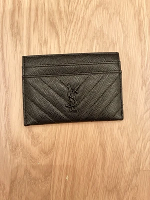 Svart YSL plånbok  - Säljer en stilren svart plånbok från YSL . Plånboken har ett quiltat mönster och flera kortfack. Framsidan pryds av den klassiska YSL-loggan i svart metall. Perfekt för dig som vill ha en lyxig och smidig accessoar i fickan.