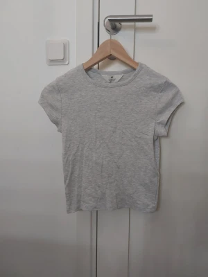 Ljusgrå basic t-shirt från H&M - En  enkel cropped och stilren ljusgrå t-shirt från H&M med rund halsringning och korta ärmar. Tillverkad i mjuk bomull som känns skön mot huden. Perfekt till jeans eller shorts för en clean look. Aldrig använt
